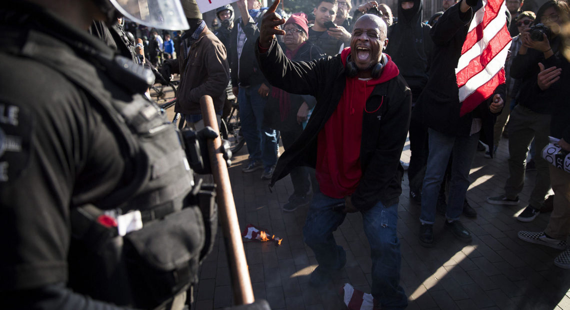 Jamal X, center, yells at police officers after lighting a portion of an American Flag on fire while protesting outside of a College Republicans rally on Saturday, Feb. 10, 2018, at Red Square on the University of Washington campus in Seattle. CREDIT: MEGAN FARMER