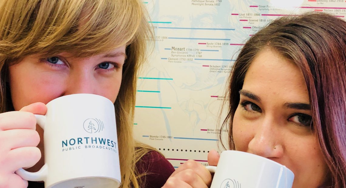 Classical music hosts Jessie Jacobs and Anjuli Dodhia drinking tea from mugs in front of a poster of musical history.