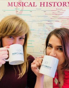 Classical music hosts Jessie Jacobs and Anjuli Dodhia drinking tea from mugs in front of a poster of musical history.