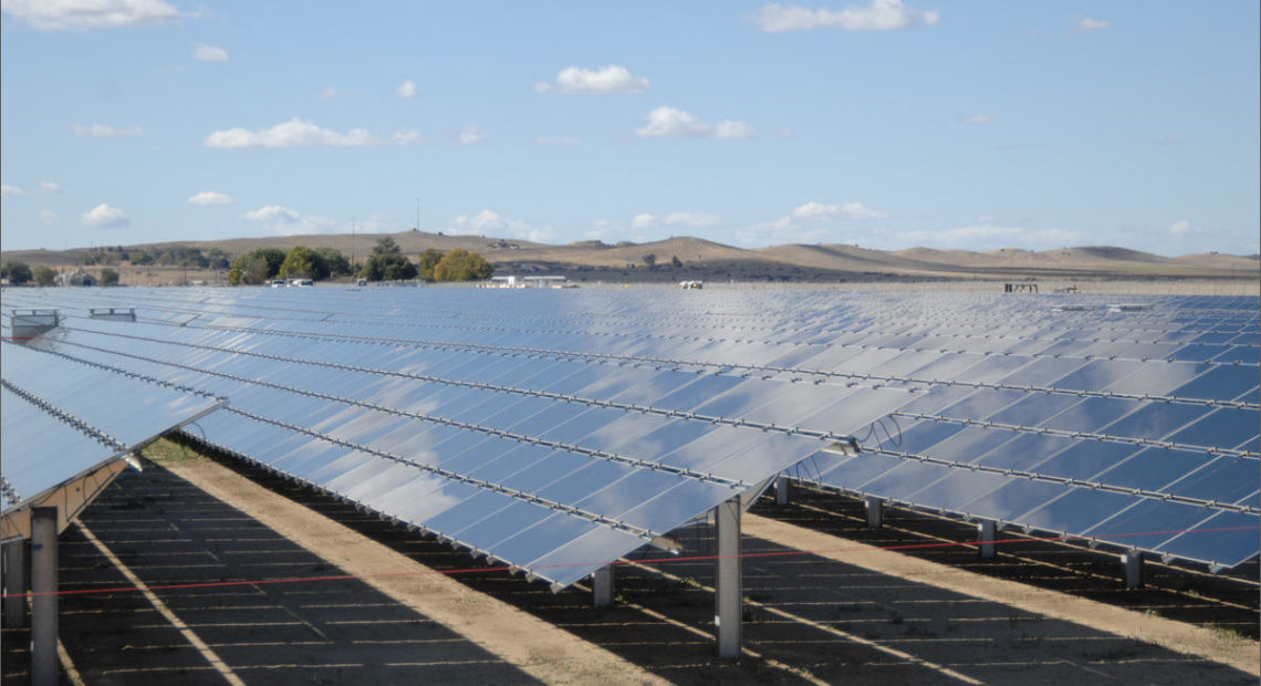 Blue-sliver solar panels in the sun. They on top of brown dirt ground. The sky is blue with little white clouds. The shadow of the solar panels are directly beneath the panels.