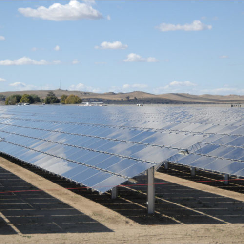 Blue-sliver solar panels in the sun. They on top of brown dirt ground. The sky is blue with little white clouds. The shadow of the solar panels are directly beneath the panels.