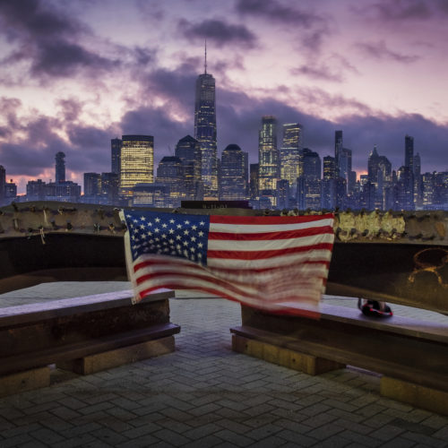 A U.S. Flag hanging from a steal girder, damaged in the Sept. 11, 2001 attacks on the World Trade Center, blows in the breeze at a memorial in Jersey City, N.J., Sept. 11, 2019 as the sun rises behind the One World Trade Center building and the re-developed area where the Twin Towers of World Trade Center once stood in New York City on the 18th anniversary of the attacks.