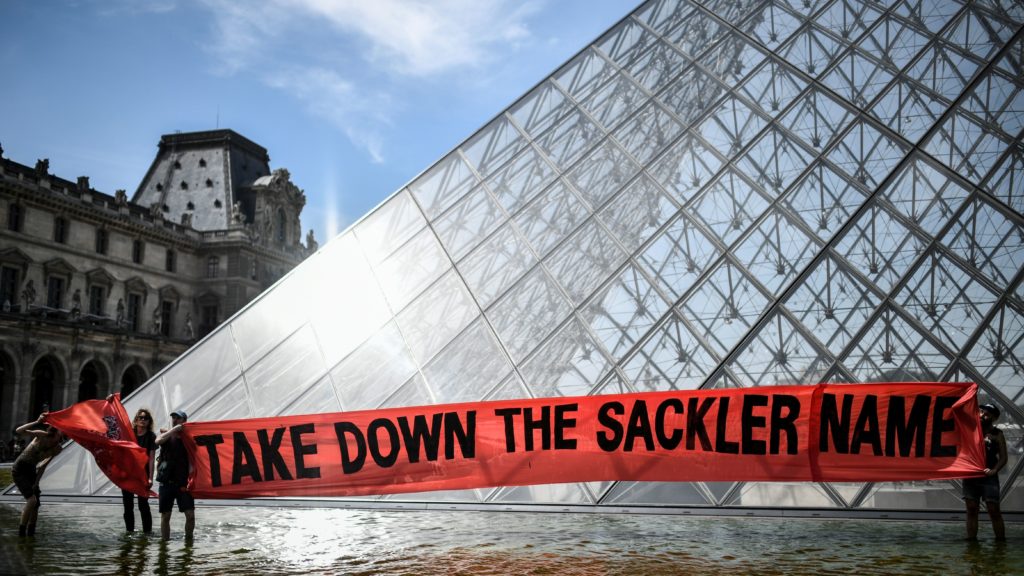Activists raise a banner reading "Take down the Sackler name" during a demonstration earlier this year in front of the Louvre Pyramid in Paris. CREDIT: Stephane de Sakutin/AFP via Getty Images