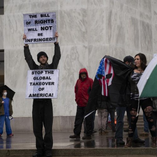 A protestor holds signs on the steps of the Oregon state capitol during a rally to reopen Oregon while health care workers counter demonstrate in the background CREDIT: Jonathan Levinson/OPB