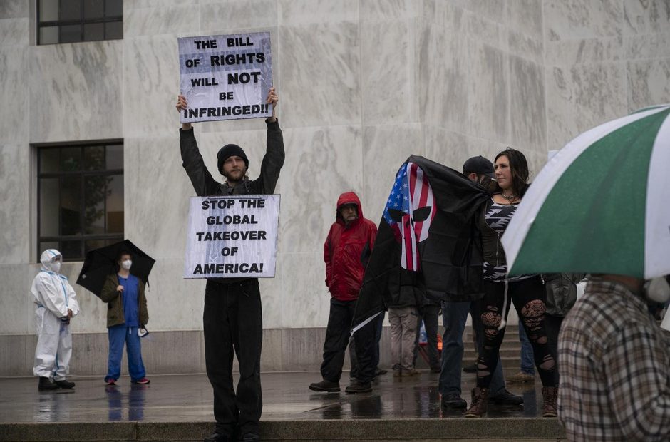 A protestor holds signs on the steps of the Oregon state capitol during a rally to reopen Oregon while health care workers counter demonstrate in the background CREDIT: Jonathan Levinson/OPB