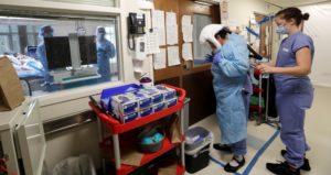 A medical worker is assisted into PPE while standing inside an area marked in blue tape, a "warm" area, before stepping into a patient's room in the COVID-19 Intensive Care Unit at Harborview Medical Center Friday, May 8, 2020, in Seattle