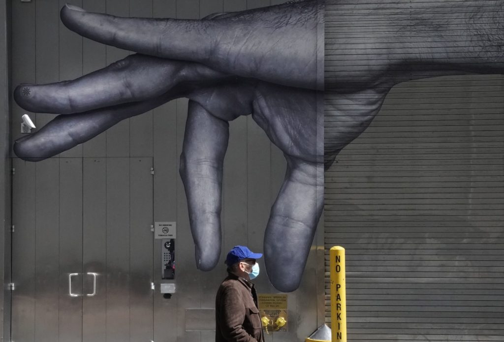 A man in a mask walks past a mural on the side of a building in New York City in April.