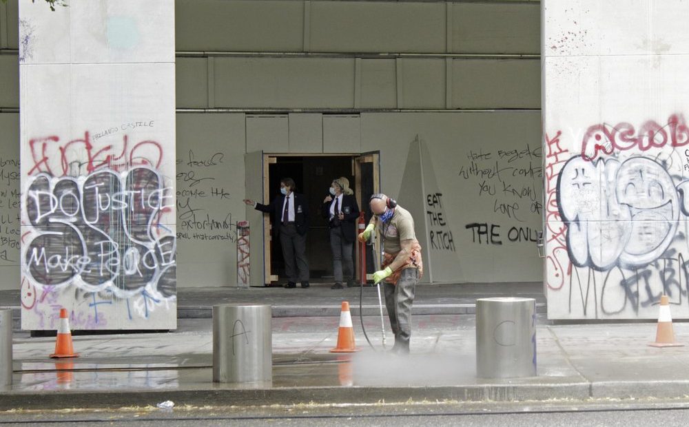 In this July 8, 2020 photo, a worker washes graffiti off the sidewalk in front of the Mark O. Hatfield Federal Courthouse in downtown Portland, as two agents with the U.S. Marshals Service emerge from the boarded-up main entrance to examine the damage. Protests and clashes with city, state, and, more recently, federal law enforcement have become a nightly occurrence in Oregon’s largest city. CREDIT: Gillian Flaccus/AP