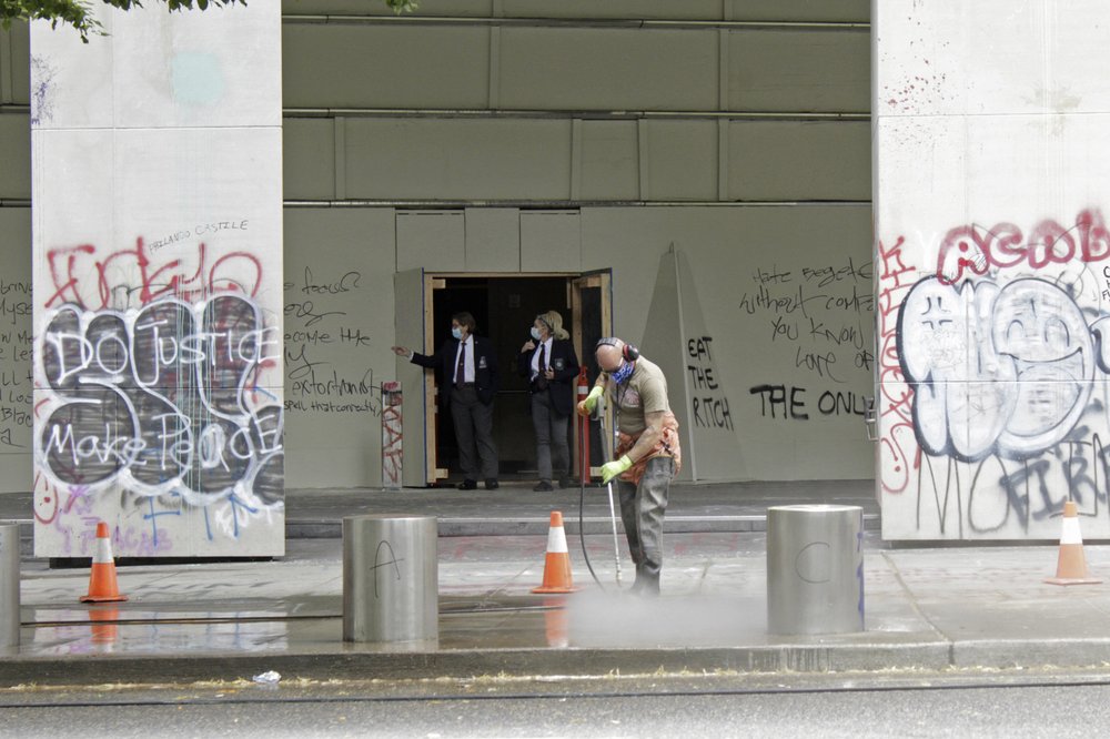 In this July 8, 2020 photo, a worker washes graffiti off the sidewalk in front of the Mark O. Hatfield Federal Courthouse in downtown Portland, as two agents with the U.S. Marshals Service emerge from the boarded-up main entrance to examine the damage. Protests and clashes with city, state, and, more recently, federal law enforcement have become a nightly occurrence in Oregon’s largest city. CREDIT: Gillian Flaccus/AP