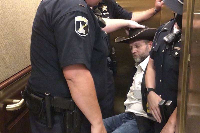 This image taken from video shows anti-government activist Ammon Bundy, rear, being wheeled into an elevator in a chair following his arrest at the Idaho Statehouse in Boise, Idaho, Tuesday, Aug. 25, 2020. Authorities arrested Bundy after he refused to leave a meeting room where a few hours earlier angry protesters forced out lawmakers. CREDIT: Keith Ridler/AP