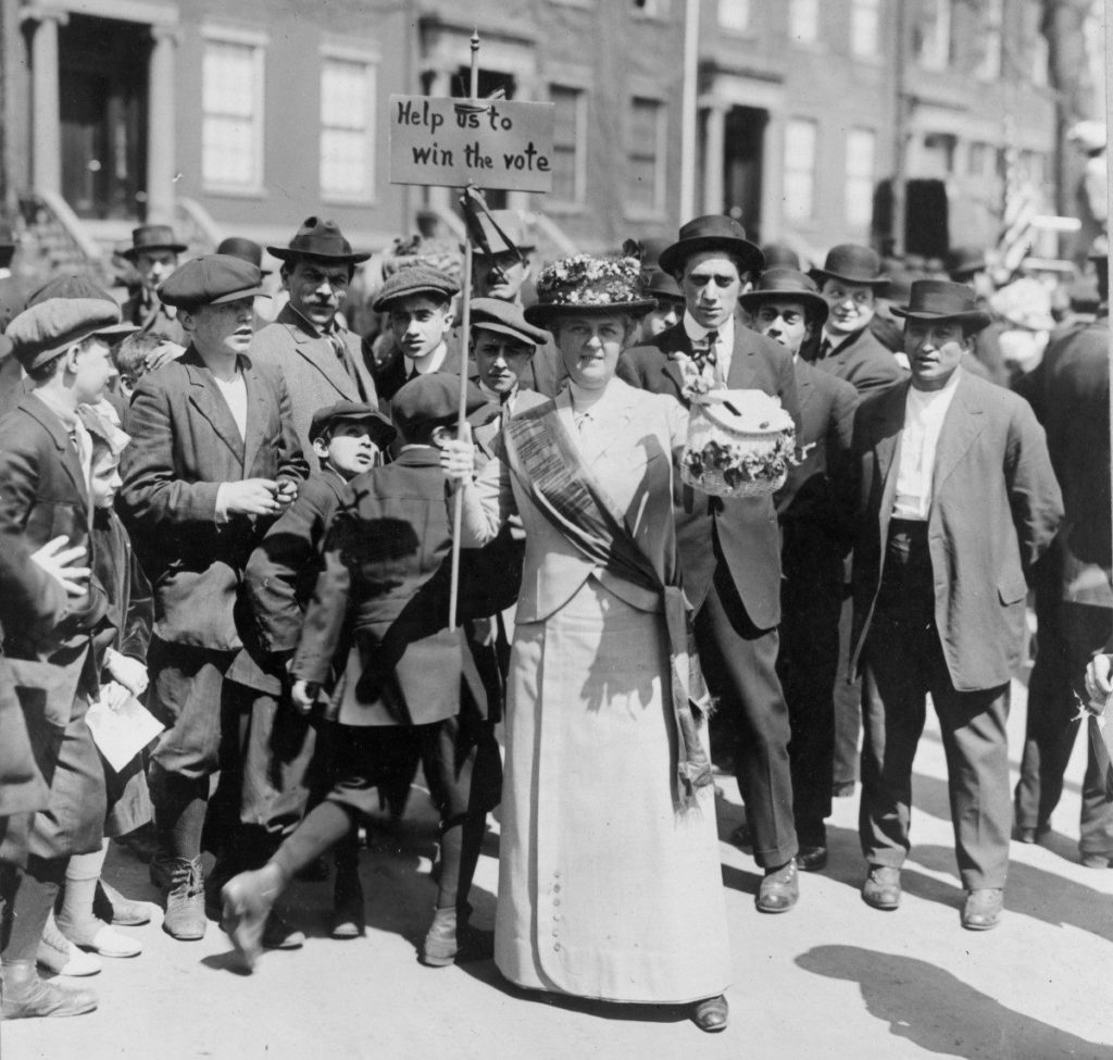 Mrs. Suffern, wearing a sash and carrying a sign that says "Help us to win the vote," surrounded by a crowd of men and boys, ca. 1914. Photo Courtesy Library of Congress/ Bain News Service