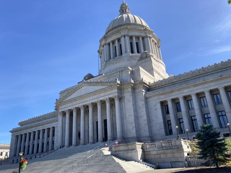 The light gray pillars, steps, and dome of the Washington State Capitol, shown at an angle in bright sunlight with a clear blue sky above. 