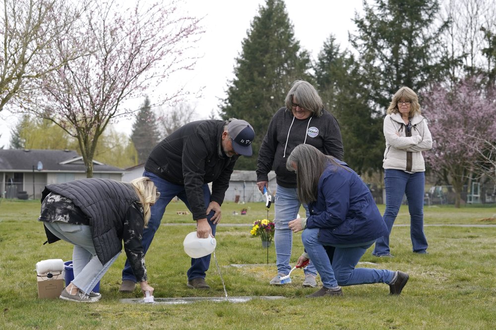 From front left, Wendy Jensen, Joe Woodmansee, Linda Holeman and Bonnie Dawson, the four children of Carole Rae Woodmansee, are joined by family friend Debbie Blazina, at right, Saturday, March 27, 2021, as they clean the headstone Carole shares with their father, Jim, who died in 2003, at Union Cemetery in Sedro-Woolley, Wash. Carole died a year ago on the same date in 2020, the day of her 81st birthday, from complications of COVID-19 after contracting it during a choir practice that sickened 53 people and killed two. CREDIT: Ted S. Warren/AP