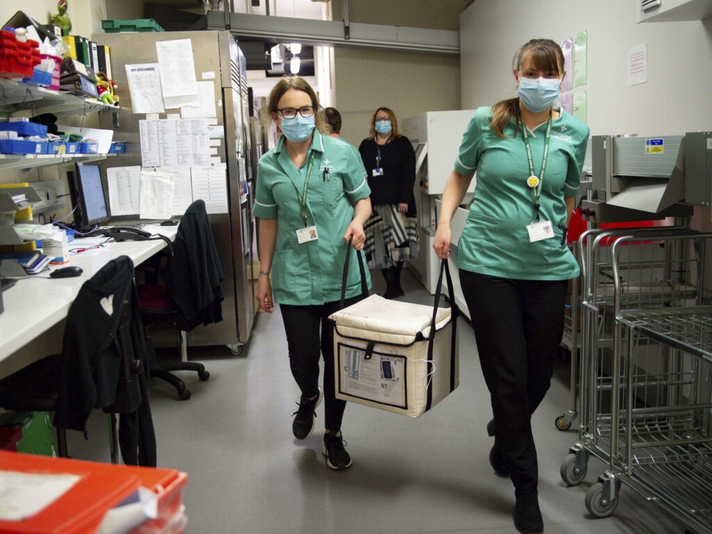 On Wednesday, pharmacists at the West Wales General Hospital in Carmarthen, Wales, transport a cool box containing the first batch of Moderna vaccines being distributed in Britain. CREDIT: Jacob King/AP