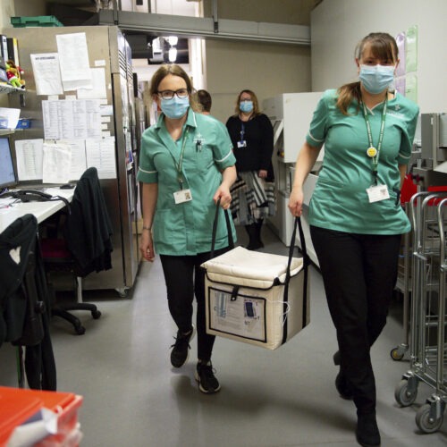 On Wednesday, pharmacists at the West Wales General Hospital in Carmarthen, Wales, transport a cool box containing the first batch of Moderna vaccines being distributed in Britain. CREDIT: Jacob King/AP