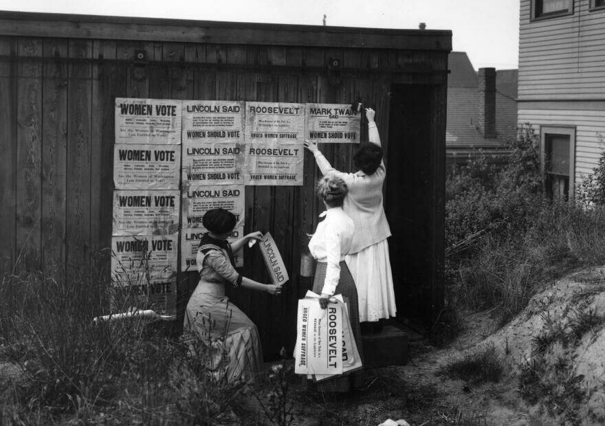 Black and white photo of three women putting up signs about Women's voting rights.