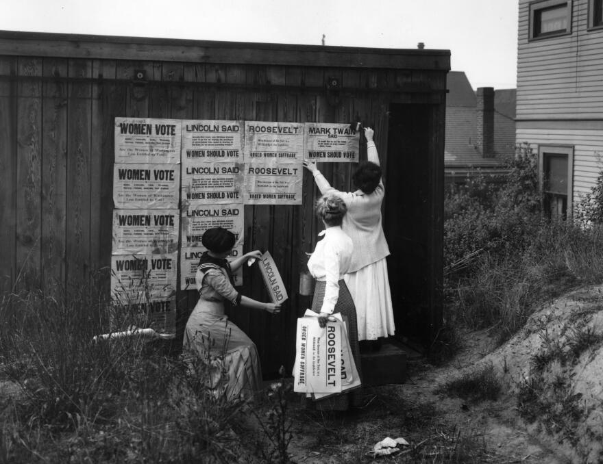 Black and white photo of three women putting up signs about Women's voting rights. 
