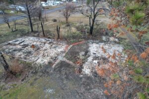 Charred ruins in grey ash sit on wet ground surrounded by trees.