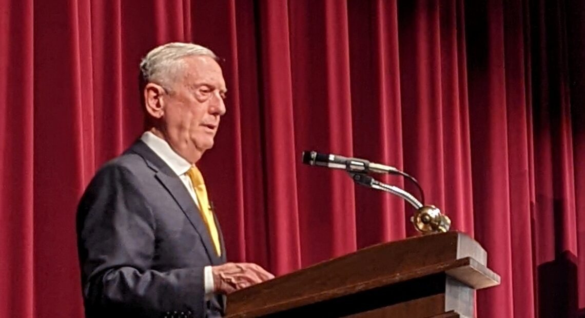 James Mattis stands at a wood podium in front of a red curtain.