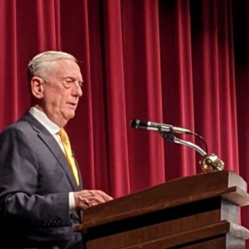 James Mattis stands at a wood podium in front of a red curtain.
