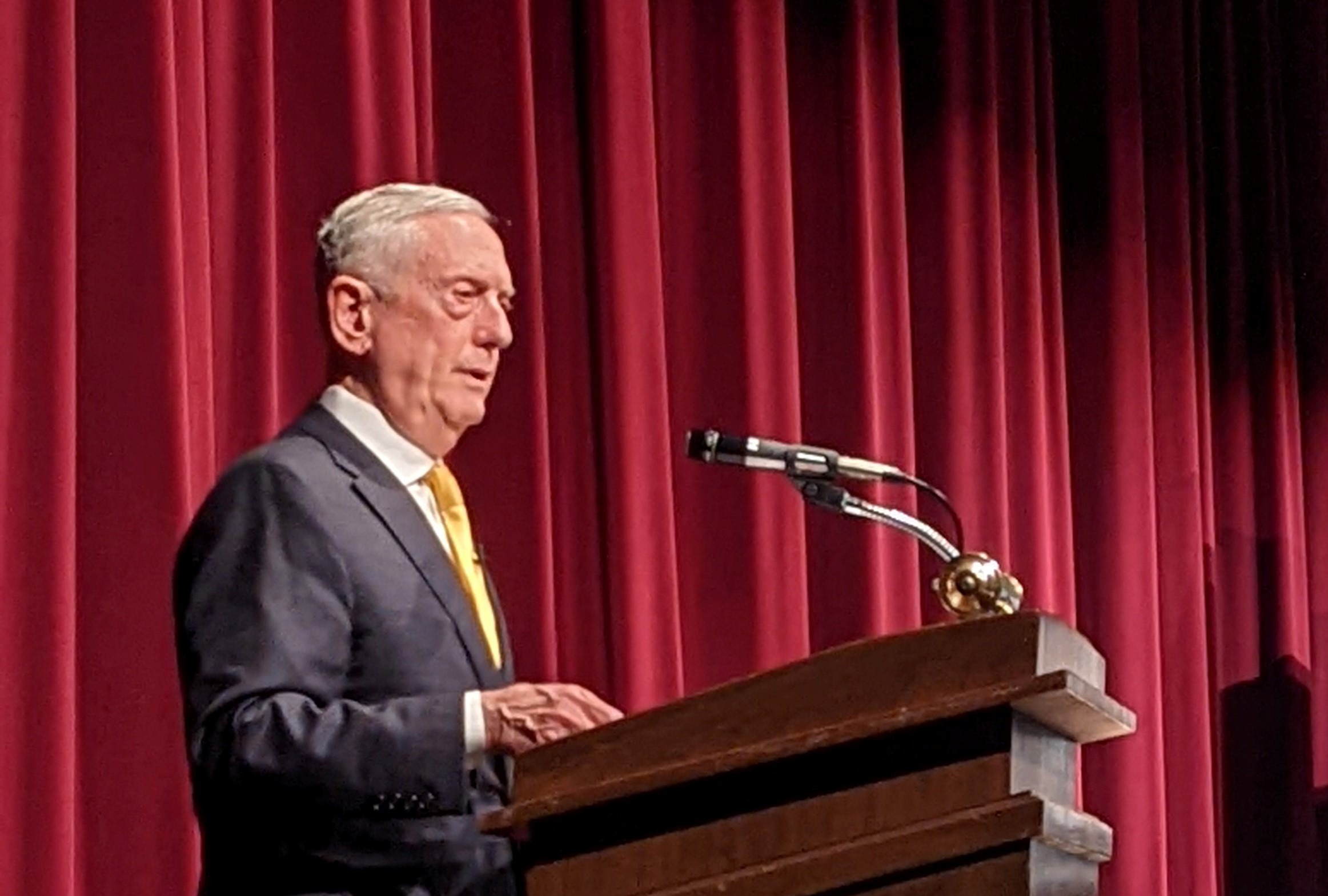 James Mattis stands at a wood podium in front of a red curtain.