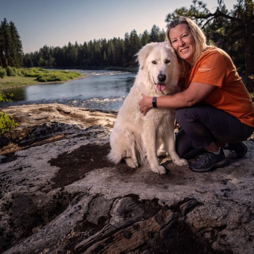 Angela Schneider sits with her big white dog atop a rock in front of a bright blue river with green trees on either side.