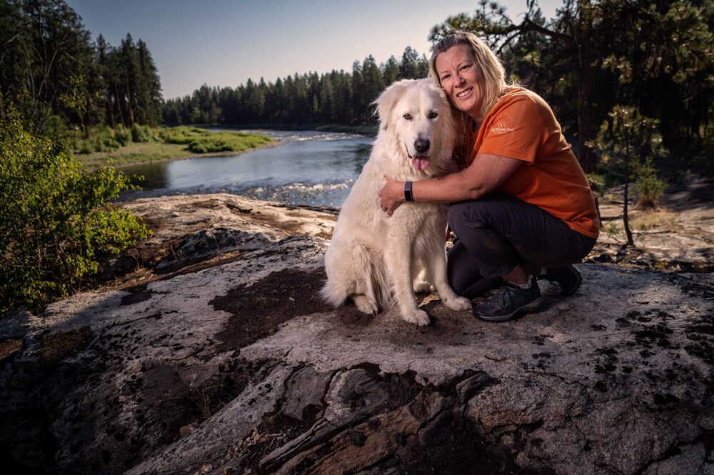 Angela Schneider sits with her big white dog atop a rock in front of a bright blue river with green trees on either side.