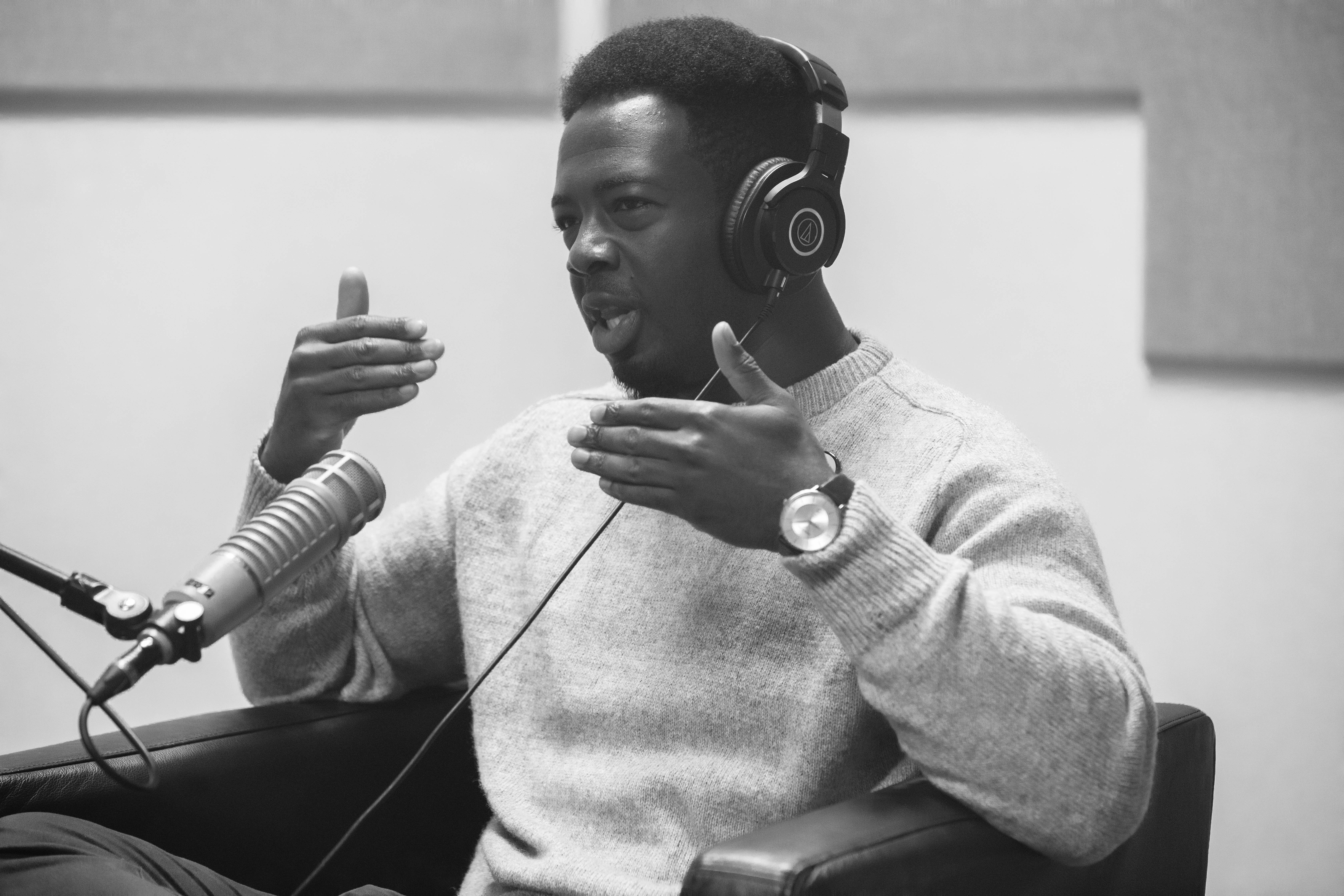 Kwabi Amoah-Forson sits in front of a microphone. His hands are in front of him as he explains something.