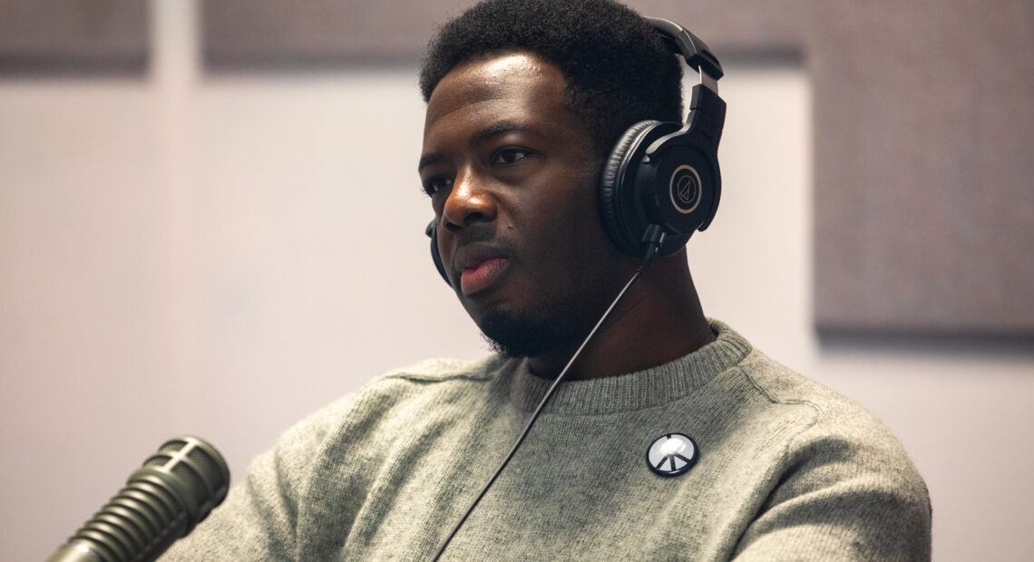 Kwabi Amoah-Forson sits in front of a microphone. He wears a pair of headphones and peace sign pin. He appears to be listening intently and with curiosity.