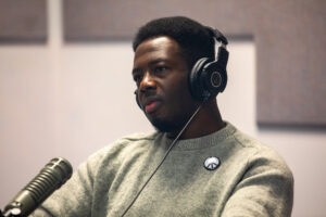 Kwabi Amoah-Forson sits in front of a microphone. He wears a pair of headphones and peace sign pin. He appears to be listening intently and with curiosity.