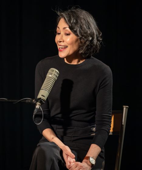 Ann Curry is looking down and smiling. She is talking into a microphone. She is wearing red lipstick and a black dress. Her hands are folded in her lap.