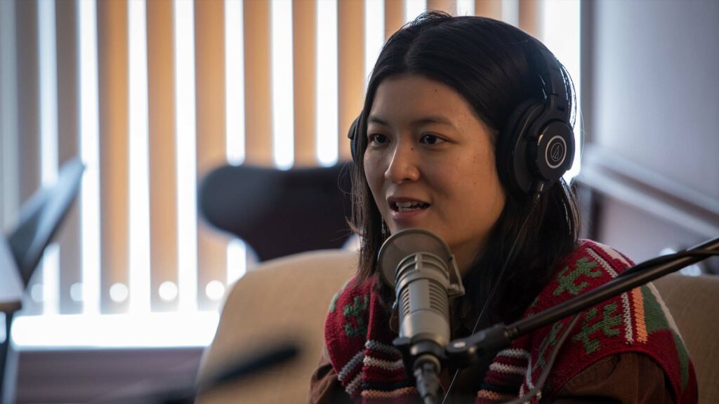 Jiemei Lin wears a pair of headphones and a crocheted sweater on a tan loveseat.