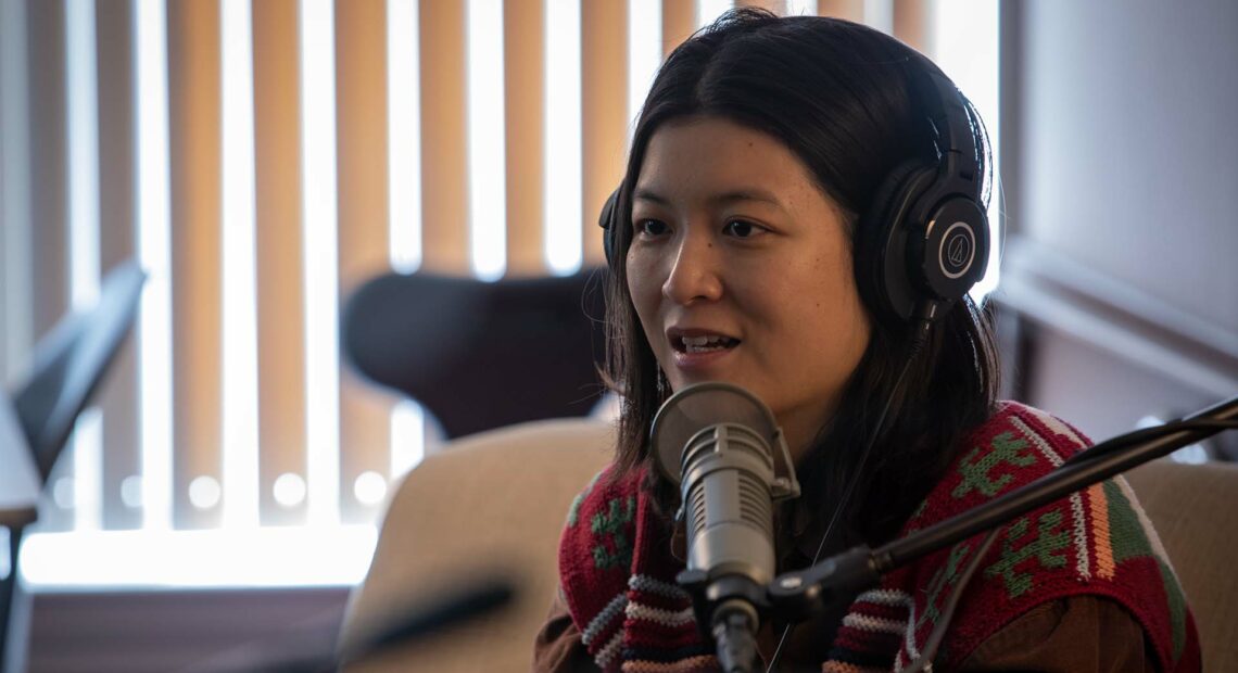 Jiemei Lin wears a pair of headphones and a crocheted sweater on a tan loveseat.