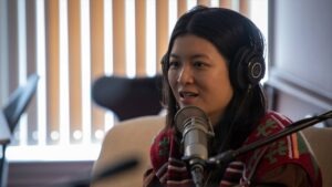 Jiemei Lin wears a pair of headphones and a crocheted sweater on a tan loveseat.