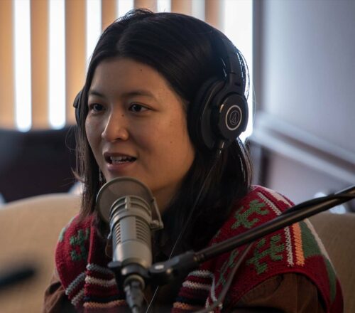 Jiemei Lin wears a pair of headphones and a crocheted sweater on a tan loveseat.