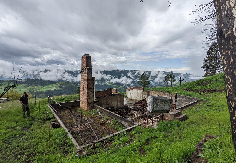 Kane Steinbruecker stands near the ash and brick ruins of a house on top of a green field.