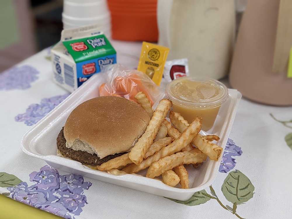A hamburger and yellow french fries sit atop a white styrofoam plate with milk nearby.