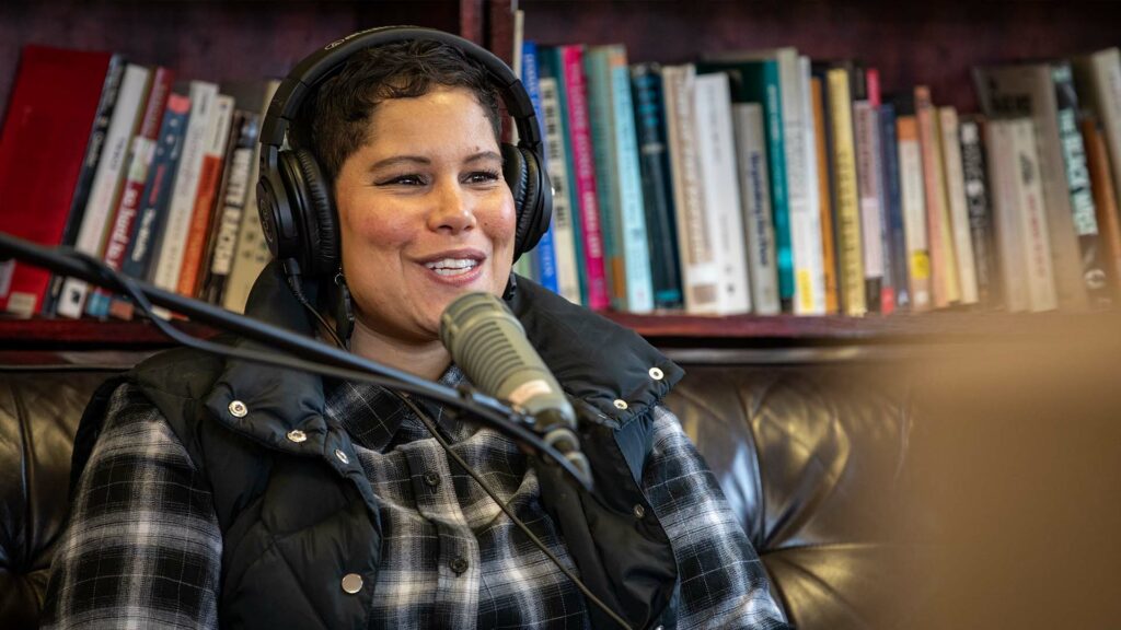 Nikkita Oliver wearing a pair of headphones, a black flannel shirt and vest smiles while sitting on a black couch in front of a bookshelf.