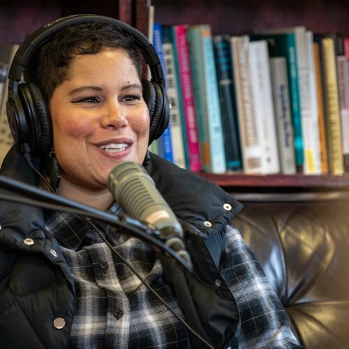 Nikkita Oliver wearing a pair of headphones, a black flannel shirt and vest smiles while sitting on a black couch in front of a bookshelf.