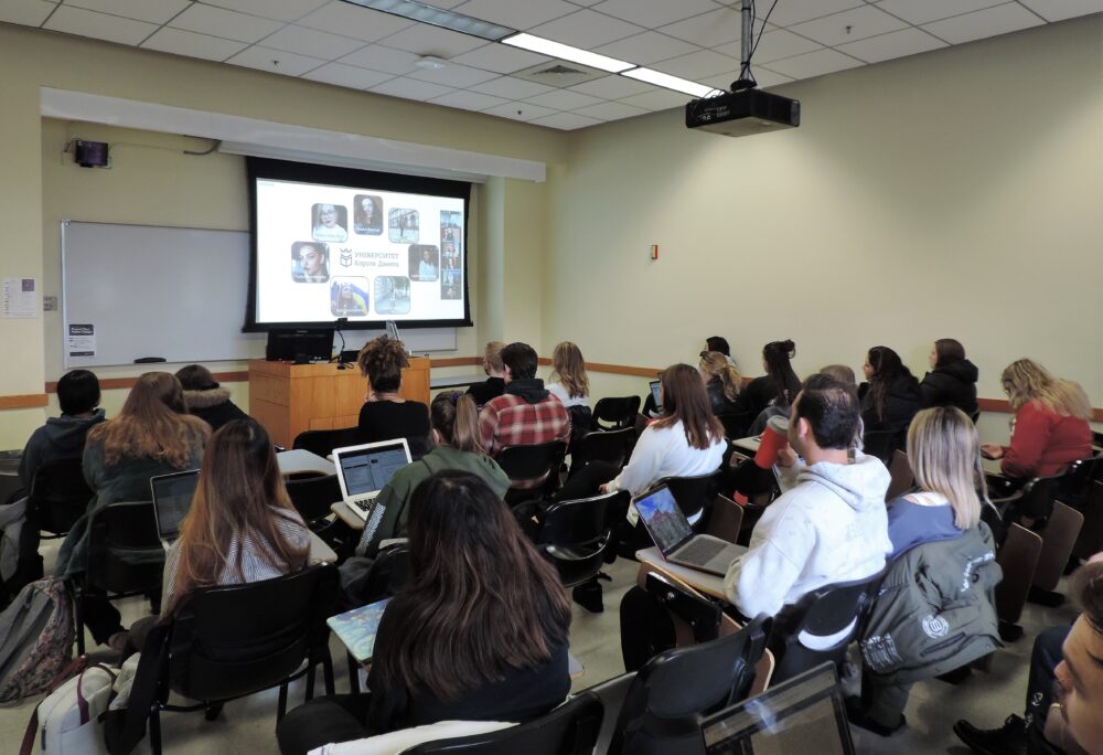 American university students sit in a classroom while the Zoom faces of Ukrainian students appear on the screen at the front of the class.