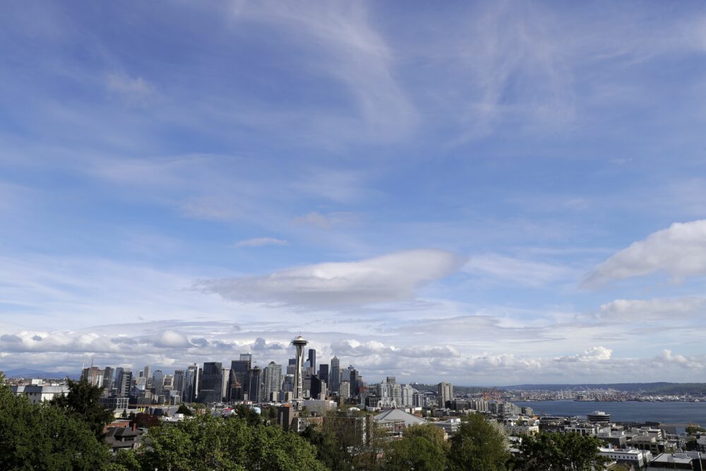 A skyline of seattle shows big skyscrapers surrounded by green foliage against a blue sky with white clouds.