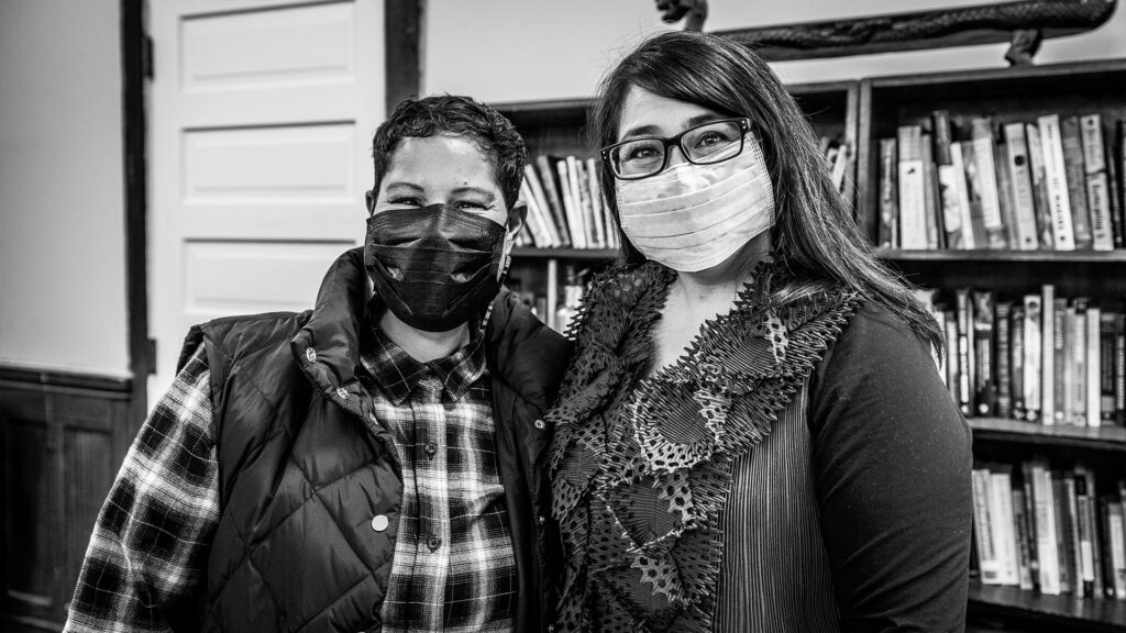Nikkita Oliver (left) and Sueann Ramella (right) stand with masks on and an arm around each other, in front of a bookcase in Washington Hall.