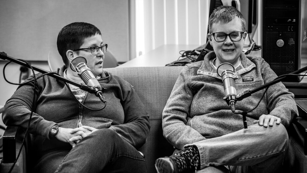 Tabitha Simmons (left) and Kathy Sprague (right) sit next to each other on a small couch, sitting with matching posture - each with their outside arm resting on the couch's armrests and their left legs crossed over their right. Kathy answers a question as Tabitha looks at her and provides additional commentary.