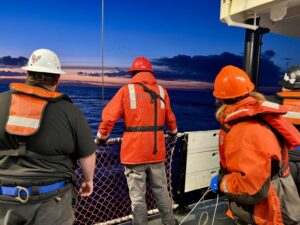 Crew members watch as a winch operator pulls a net up from the ocean.