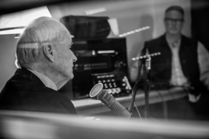 Chris, positioned on the left side of the image, is facing away from the camera, talking into his microphone. A NWPB employee is in the background of the image on the right side. There is a glare over the photograph because it was taken from outside the recording booth.