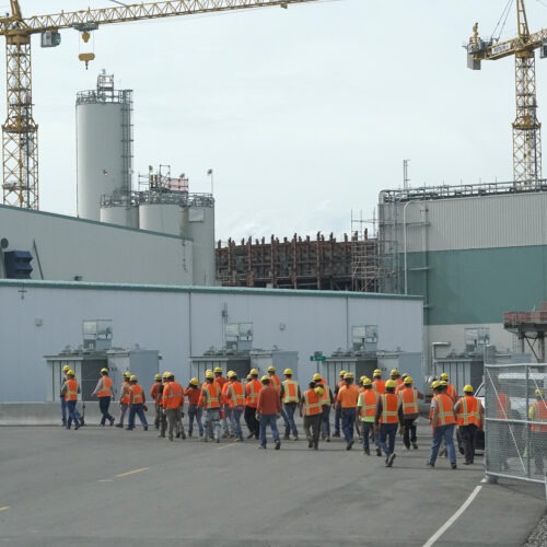 Wokers with orange vests and yellow hard hats make their way to a large grey nuclear waste facility.