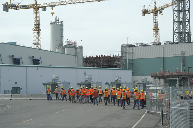 Wokers with orange vests and yellow hard hats make their way to a large grey nuclear waste facility.