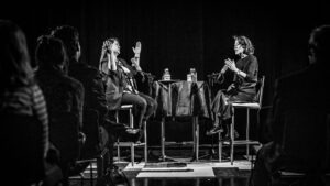 Centered in the photo's middle-ground are Sueann and Ann, sitting opposite each other at a high table. Sueann has her arms raised as she reacts to Ann's response. Ann is gesturing with her hands as she speaks. Blurred in the foreground are members of the live audience.