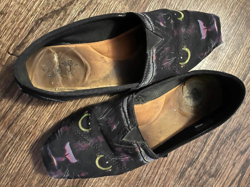 Black loafers with a cat's face on the toe side of the shoe sit on a wooden floor. The black shoes look worn.