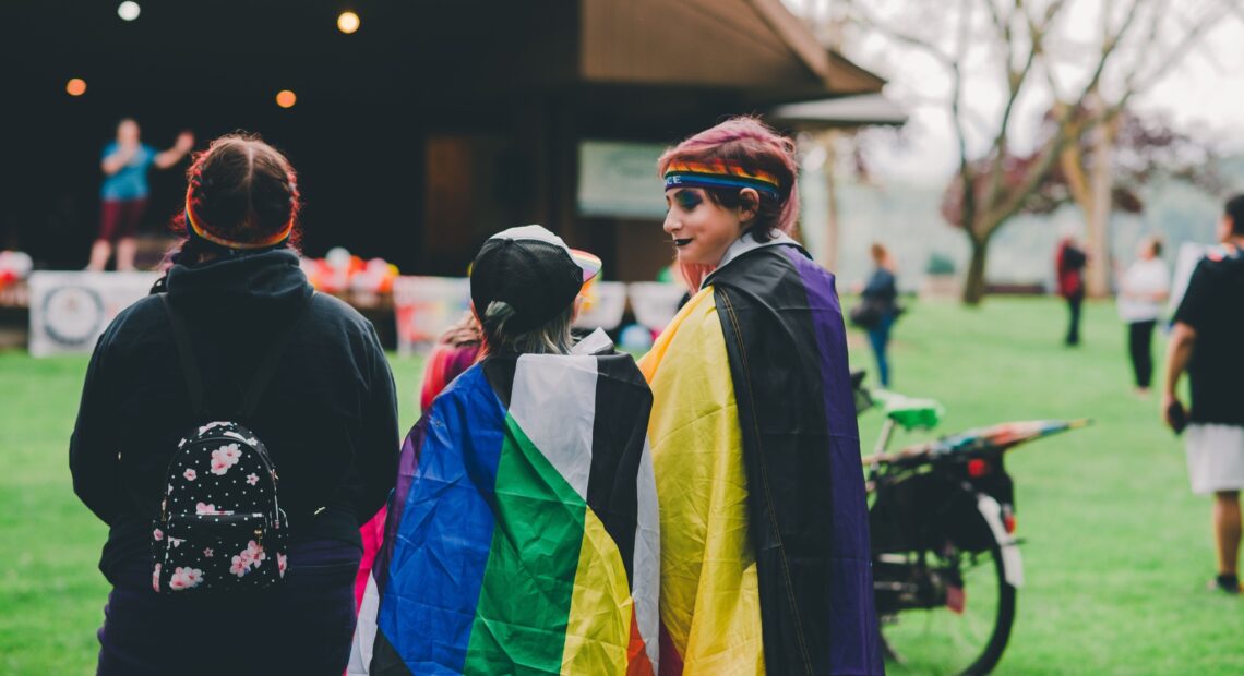 Three people stand together in a park facing a stage. One wears black lipstick, one other wears a pride flag draped around their shoulders.